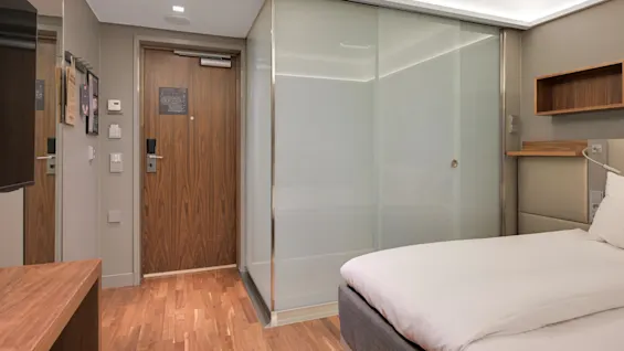 En trädörr i ett hotellrum är stängd, bredvid frostade glasväggar. En snyggt bäddad säng är delvis synlig på ett trägolv. Dämpad belysning skapar en modern och mysig atmosfär.
