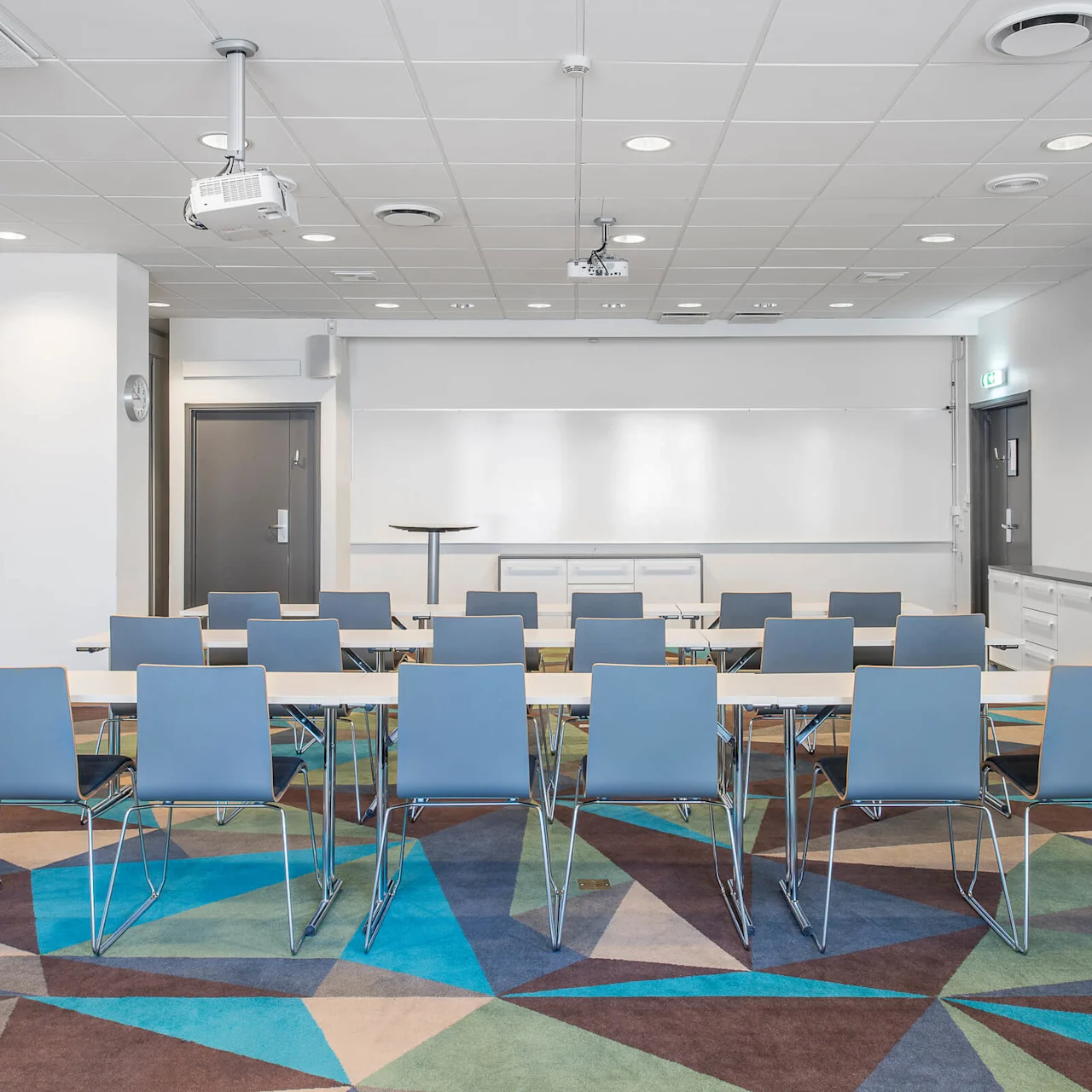Rader med blå stolar vänder sig mot en whiteboard i ett modernt konferensrum med geometriskt mönstrad matta och takprojektorer. En dörr och en utgångsskylt syns till höger.