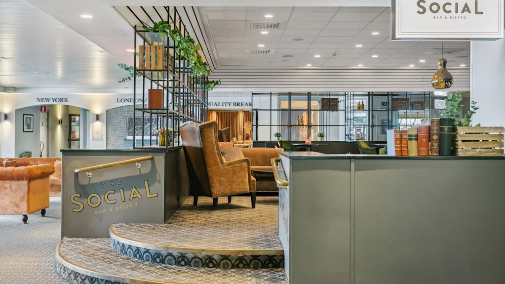 Et siddeområde med lænestole og en stilfuld metalopdeler fyldt med planter og bøger i en bistro-indstilling. Skiltet lyder: "THE SOCIAL BAR & BISTRO”.