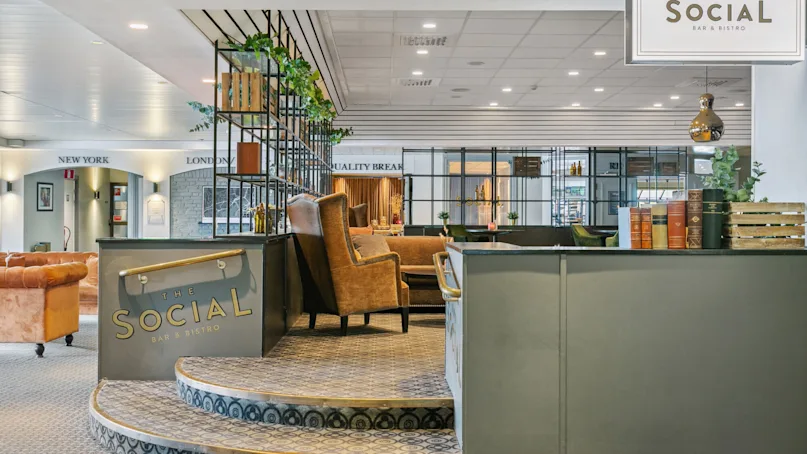A seating area with armchairs and a stylish metal divider filled with plants and books in a bistro environment. Sign reads: “THE SOCIAL BAR & BISTRO”.