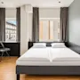 Modern hotel room featuring a double bed, light walls, wooden flooring, and two windows letting in daylight.