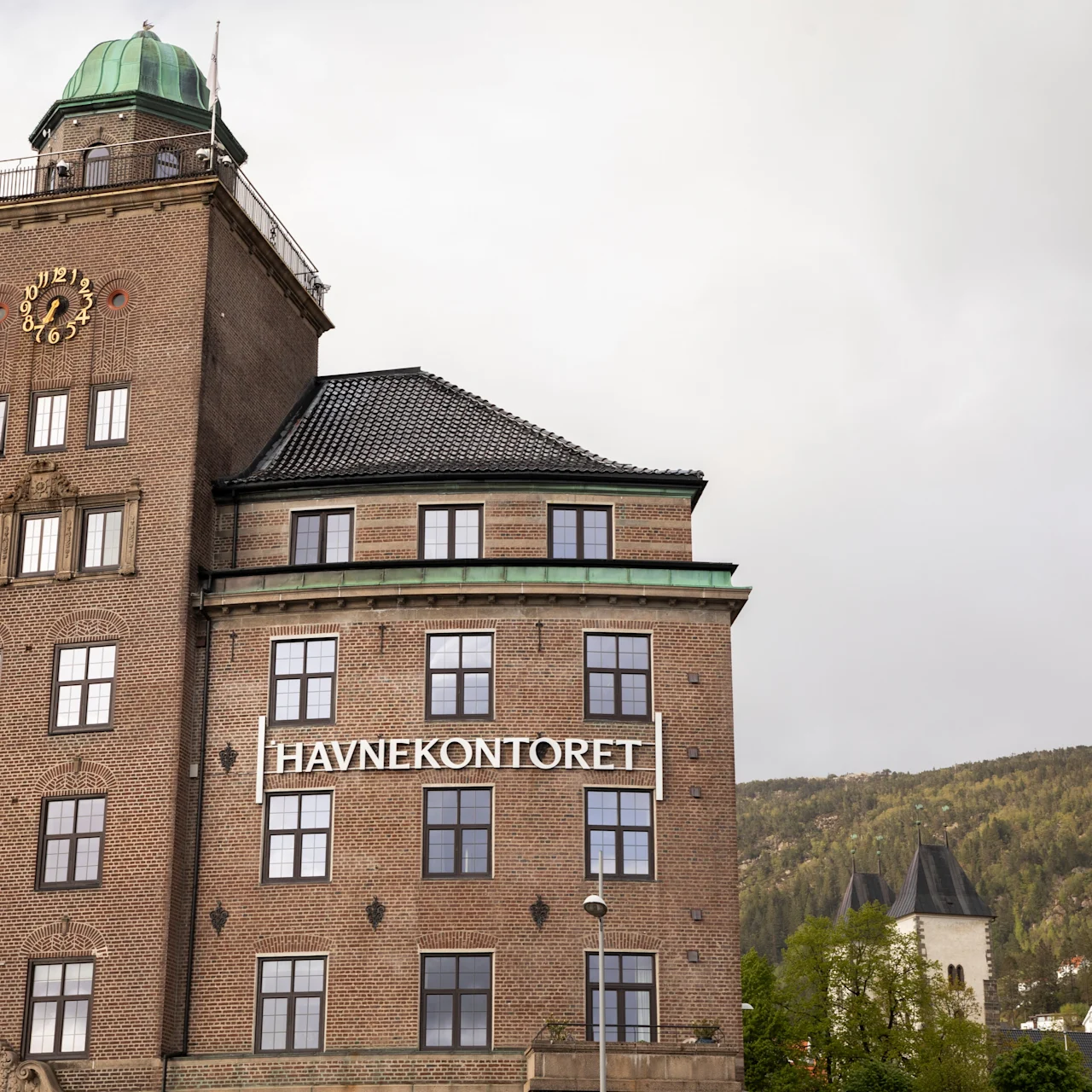 A large brick building with a clock tower displays the text "HAVNEKONTORET" on its facade. It is set against a backdrop of wooded hills and cloudy sky.