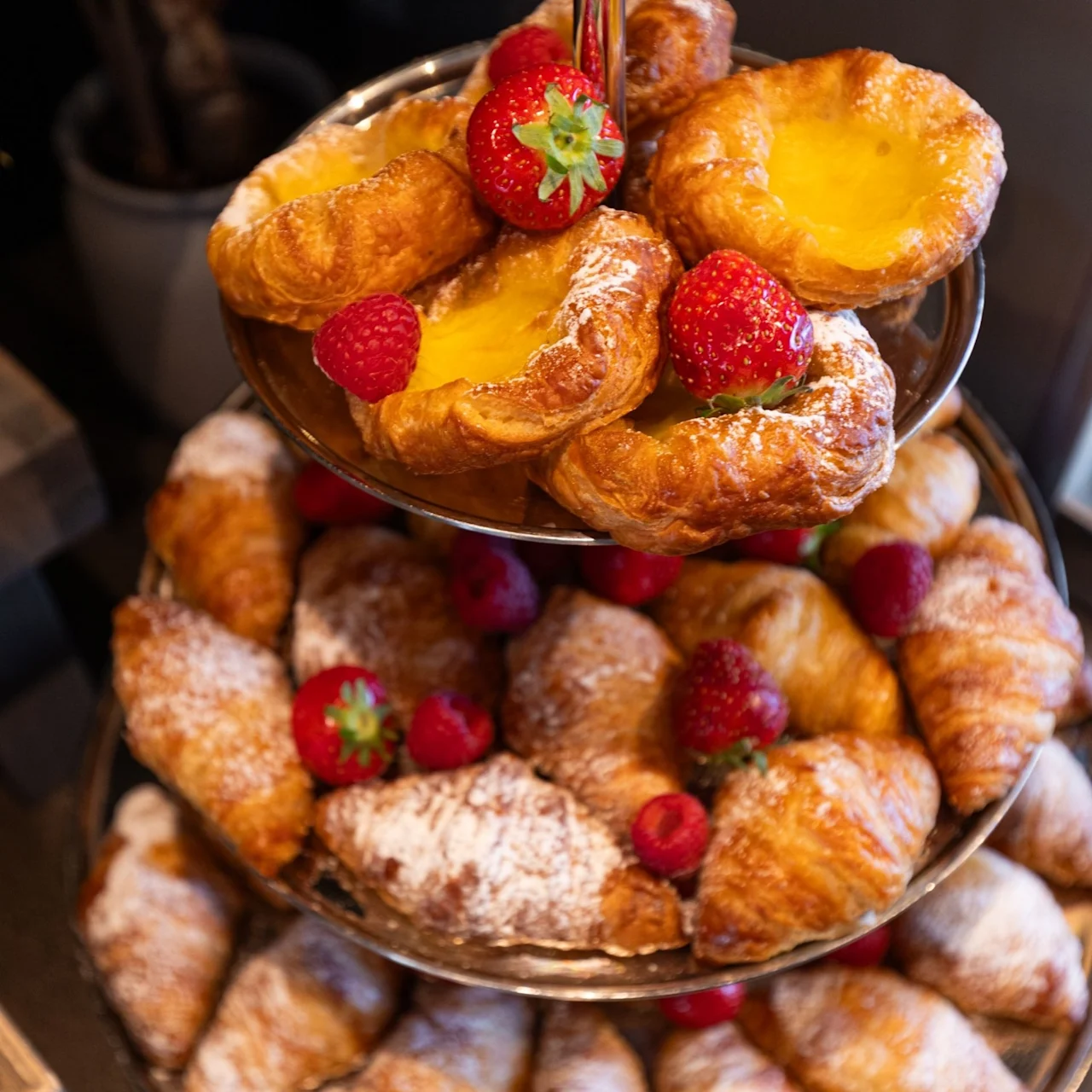 Pastries on a tiered stand, with croissants and lemon tarts topped with strawberries and raspberries, arranged in a decorative display on a table.