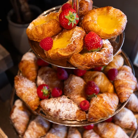 Bakverk på et etasjefat, med croissanter og sitronterter toppet med jordbær og bringebær, dekorativt arrangert på et bord.