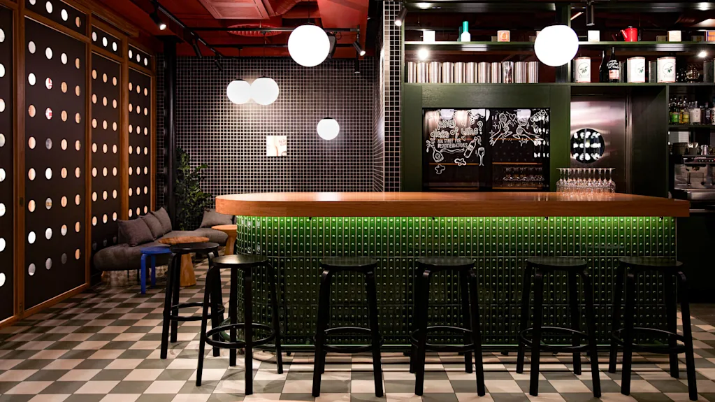 Bar with a wooden counter lined by black stools, situated in a modern lounge with checkered floor and patterned walls. Circular light fixtures hang from the red ceiling.