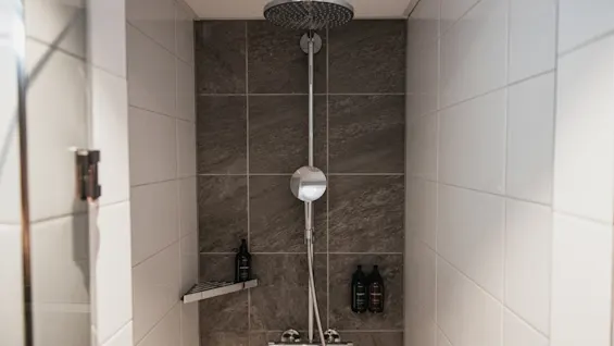 En moderne dusj med regndusjhode og toalettartikler, mot hvite og mørkegrå flislagte vegger.