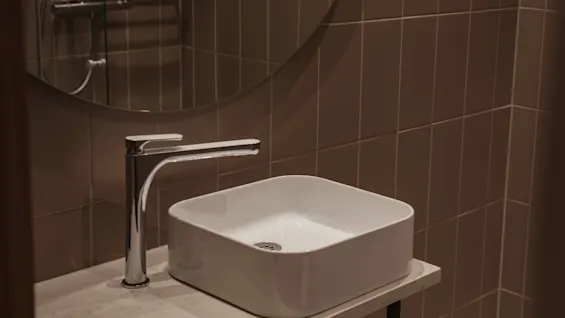 A square white sink with a sleek silver faucet sits on a marble countertop in a bathroom with brown tiled walls, beneath a circular mirror.