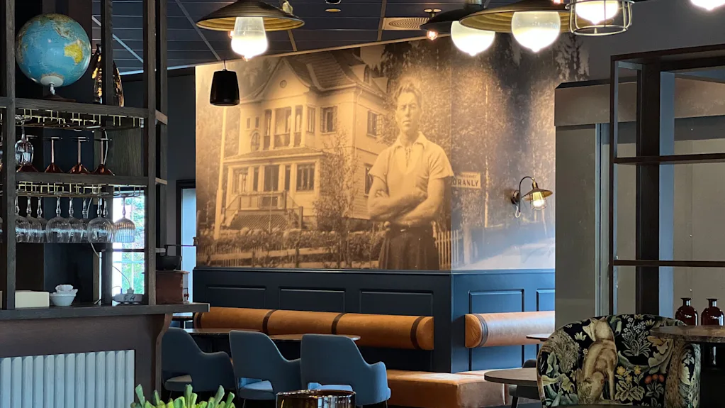 A wall displays a large vintage photograph of a man standing in front of a house. The café interior includes hanging lights, a globe, and wooden furniture.