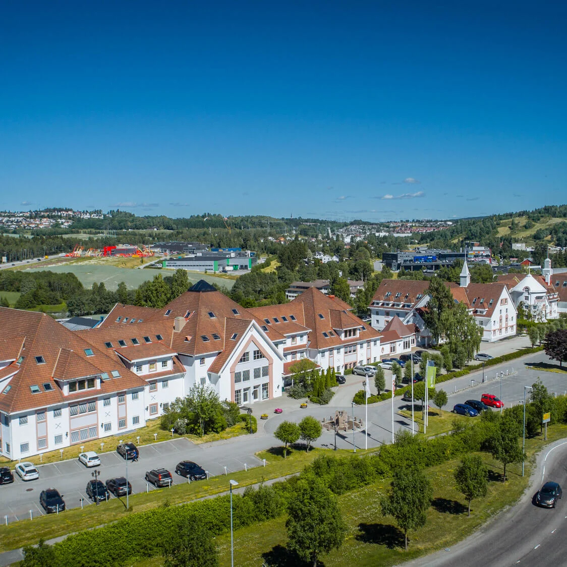 En stor bygning med røde tage står i et anlagt område omgivet af parkerede biler, tilstødende veje og fjerne bolig- og industribygninger mod grønt, kuperet terræn under en blå himmel.