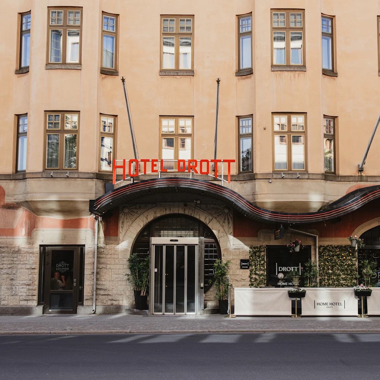 Byggefasade viser "HOTEL DROTT" i rød neonlys, med buet inngang flankert av planter. Området inkluderer vinduer og skilt for "DROTT LOUNGE" og "DROTT BAR".