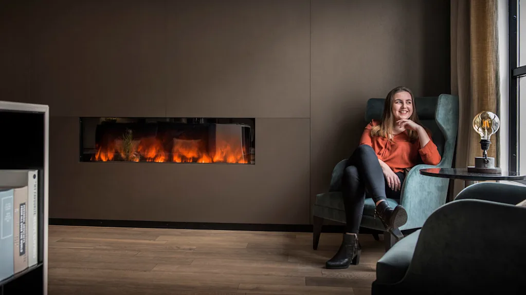 A woman sits smiling in a high-back chair beside a wall-mounted fireplace in a cozy room with brown walls and wooden floors.