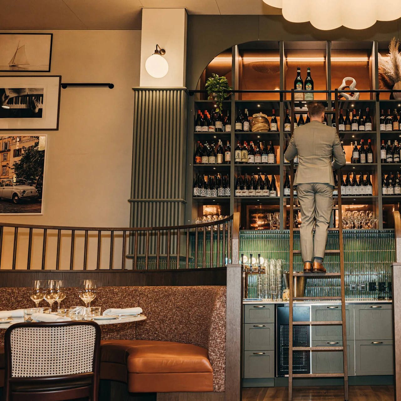 A man stands on a ladder, selecting wine bottles from a tall, well-organized shelf in an elegant restaurant with cushioned seating, framed photographs, and soft lighting.