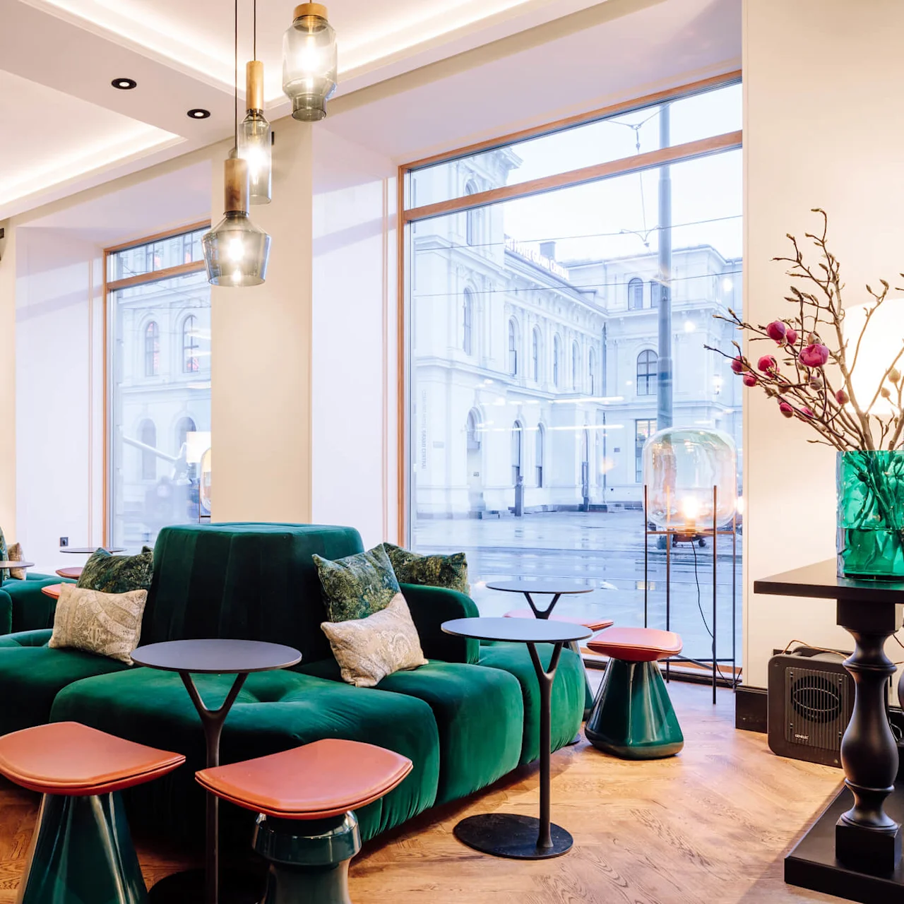 Luxurious green velvet sofa sits with decorative pillows, surrounded by small wooden tables and stools in a bright, modern lounge with large windows and hanging pendant lights.