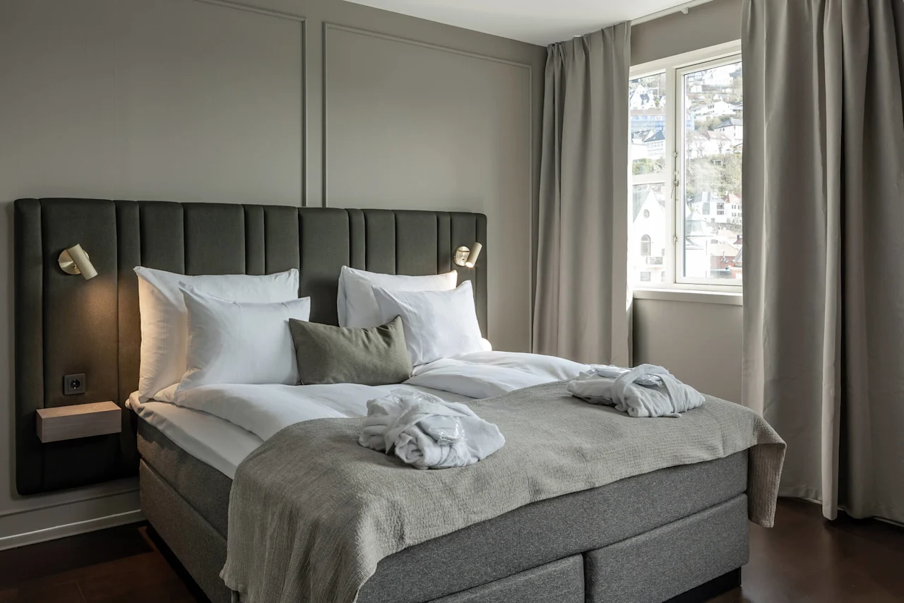 En nydelig oppredd seng med hvite puter og badekåper står mot en grå panelvegg. Myk belysning og gardiner rammer inn et vindu med utsikt over hus.