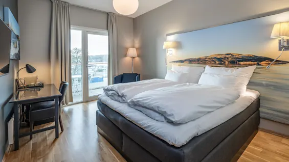 En pænt reddet seng med hvidt sengetøj står i et moderne soveværelse, ved siden af et skrivebord med en lampe, med udsigt over en balkon gennem glasdøre.