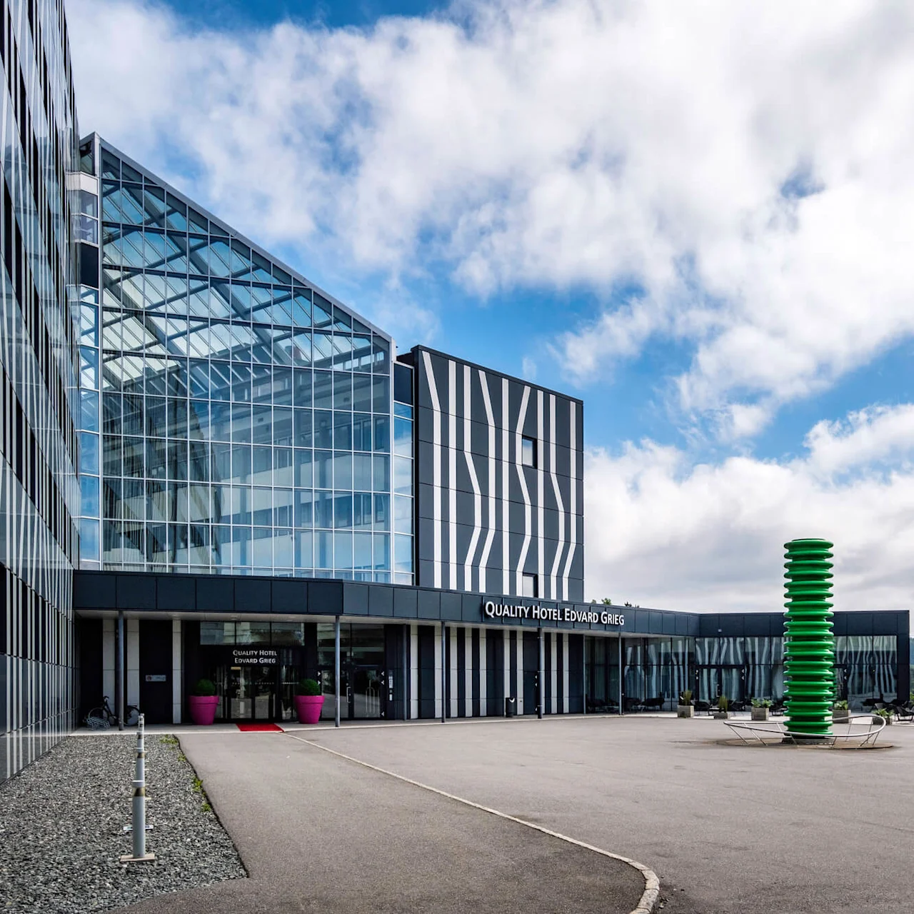 En moderne hotellbygning med teksten "QUALITY HOTEL EDVARD GRIEG" har glassfasader med abstrakte mønstre. Flaggstenger og en grønn skulptur står utenfor mot en delvis skyet himmel.