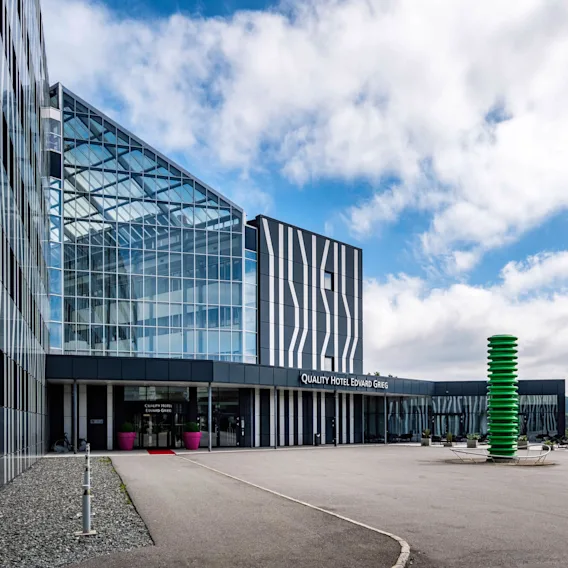 En moderne hotellbygning med teksten "QUALITY HOTEL EDVARD GRIEG" har glassfasader med abstrakte mønstre. Flaggstenger og en grønn skulptur står utenfor mot en delvis skyet himmel.