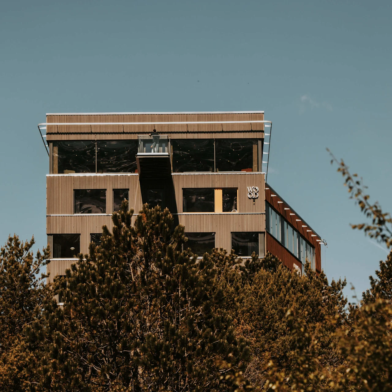 A modern building with "WOOD" text visible, surrounded by green trees against a clear blue sky.