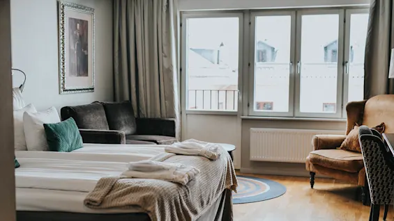Et hyggeligt og veloplyst soveværelse med en pænt redt seng, indrammet billede ovenfor, en sofa, lænestole, samt udsigt gennem franske vinduer.