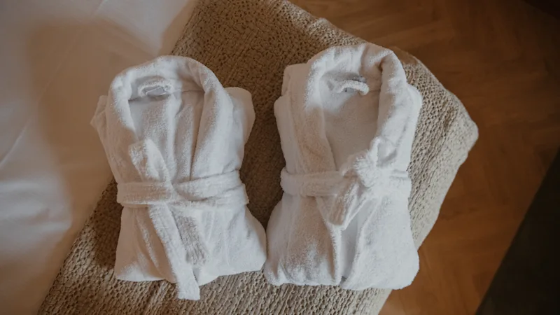To bløde hvide badekåber foldet på en seng, der inviterer gæster til at slappe af.
