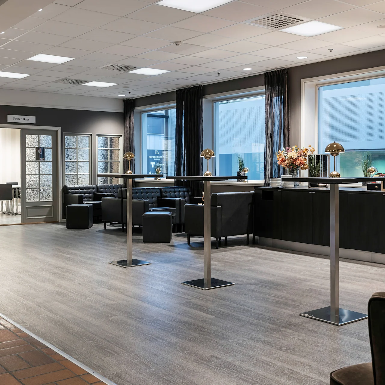 A lounge with high tables and chairs is set against large windows. Comfortable seating and decorative plants enhance the relaxed atmosphere. A sign reads "Petter Dass" above a conference room.