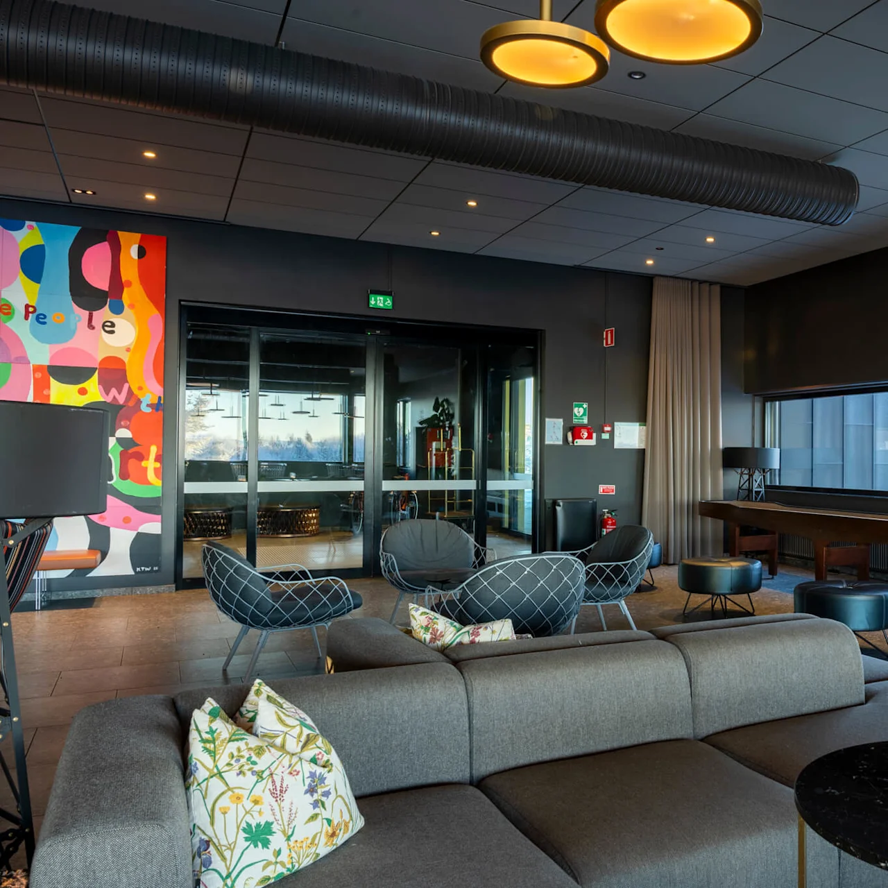 Modern lounge med grå soffor och färgglad konst på väggarna. Stämningsbelysning, stora fönster visar en utomhusvy. Skylt på konstverket lyder "people" med abstrakta mönster.