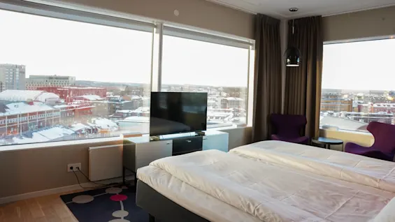 Et hotelværelse med en seng, der vender mod et fjernsyn, med udsigt over et sneklædt bybillede gennem store vinduer. Tekst på en fjern bygning: "Comfort Hotel".