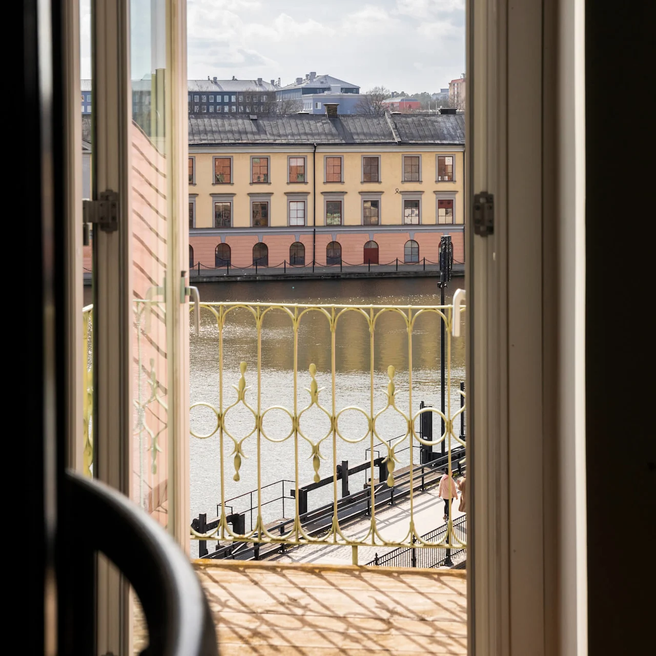 Öppet fönster leder till en balkong med utsmyckat räcke, med utsikt över en lugn flod och pastellfärgad byggnad under en klarblå himmel.