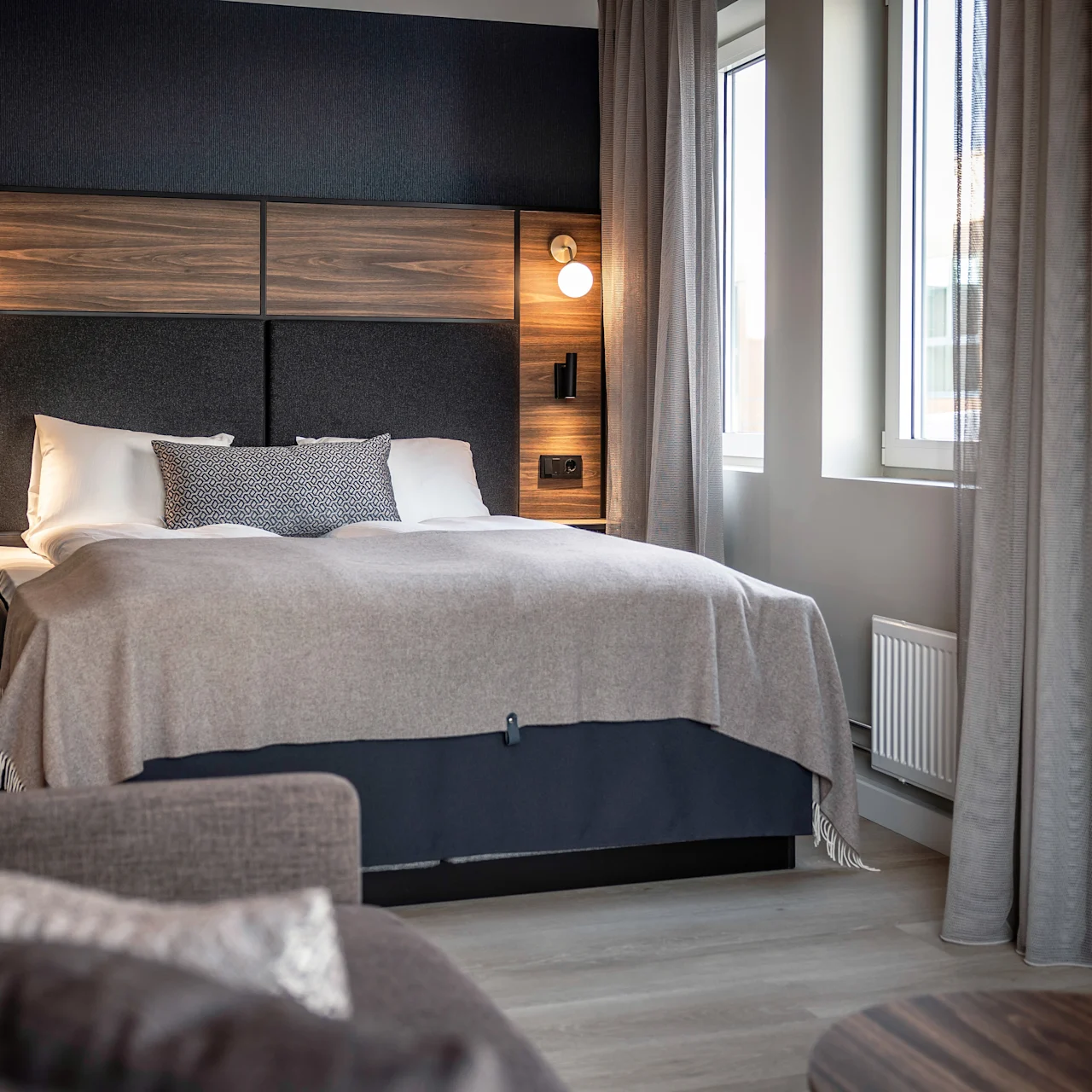 Säng med trähuvudgavel och geometriska kuddar, under varm belysning. Omgiven av en mjuk soffa, fönster med gardiner och neutrala väggar, vilket skapar en mysig, modern sovrumsatmosfär.
