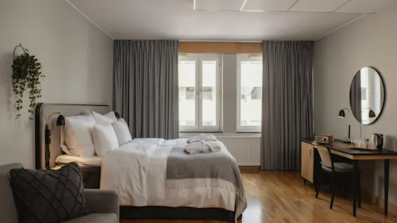 En pent oppredd seng med puter og et sammenbrettet teppe står i et romslig soverom, som også har et skrivebord og en stol ved et vindu med myke, grå gardiner.