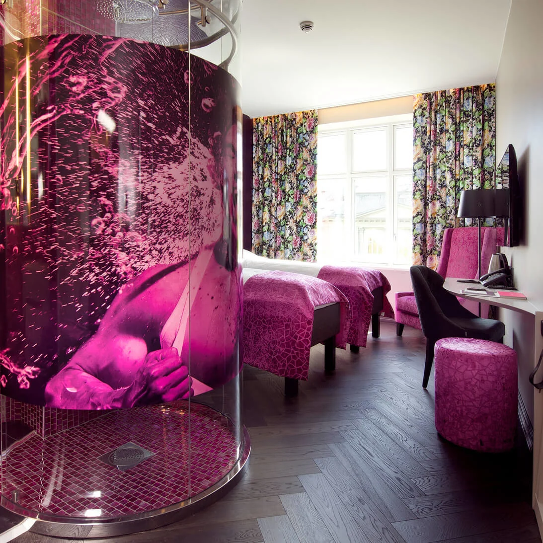 A vibrant room features a pink shower with artistic designs, adjacent to a modern bed with floral curtains. A desk, chair, and textured decor complete the lively atmosphere. Text: "NA."