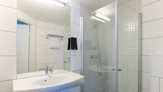 A modern bathroom with a sink, mirror, shower, and towels, featuring white tiles and clean design.