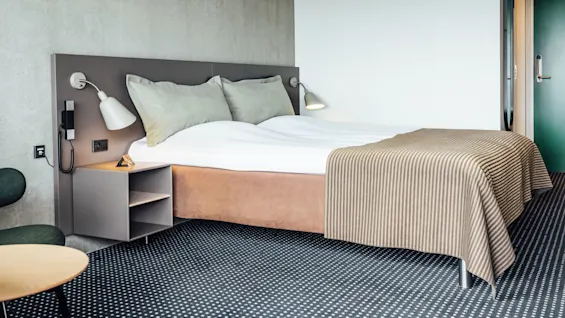 Seng med hvidt sengetøj og stribet tæppe placeret i et moderne, minimalistisk hotelværelse; har grå vægge, en grøn dør og et hyggeligt sidebord.  