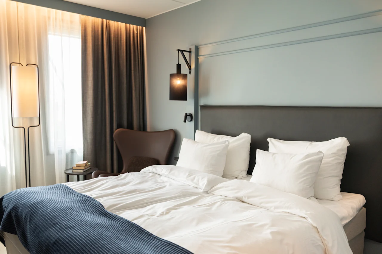 Et koselig hotellrom med en seng med hvitt sengetøy og et blått pledd, en lenestol og varm belysning.