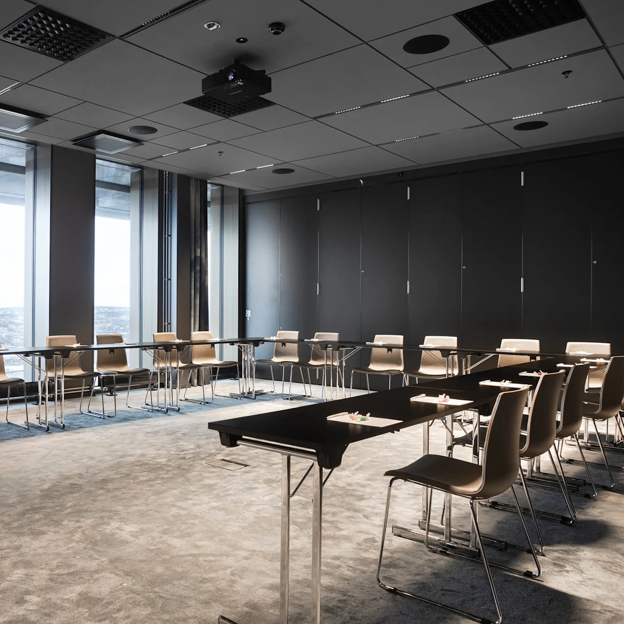 Konferensrum med arrangerade stolar runt bord, vy över stadsbild genom stora fönster. Modern takbelysning, projektor centrerad ovanför och neutralfärgad matta.