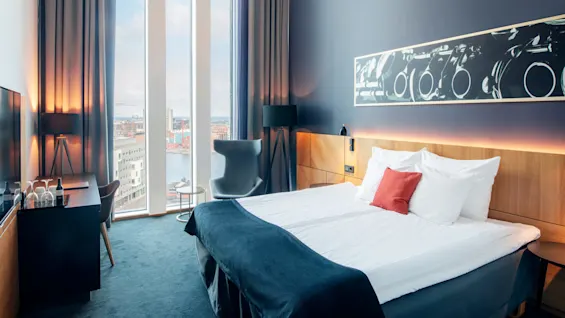 En stor seng med hvitt sengetøy og en rød pute står i et moderne hotellrom med gulv-til-tak-vinduer og byutsikt. Et skrivebord og en stol er i nærheten.
