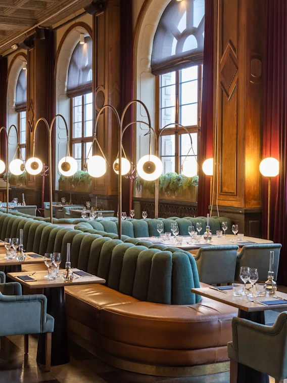 Bord och fåtöljer arrangerade i en elegant restaurang, upplysta av stilfulla hänglampor, med stora valvformade fönster längs väggen, vilket ger en sofistikerad och stämningsfull matmiljö.
