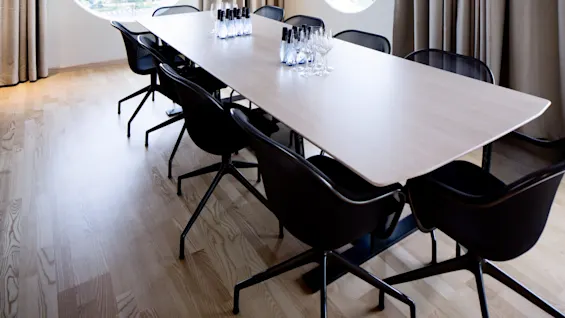 A long conference table is set with water bottles and empty glasses, surrounded by chairs. Two large pendant lights hang above. Circular windows with curtains provide natural light.