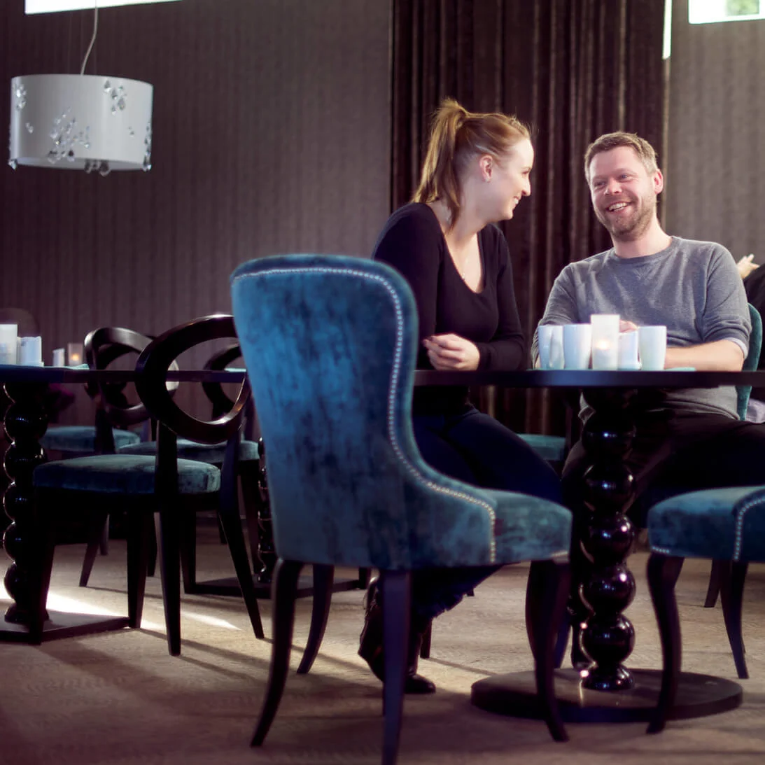 Two people are laughing and talking over coffee in a stylish café with plush blue chairs.