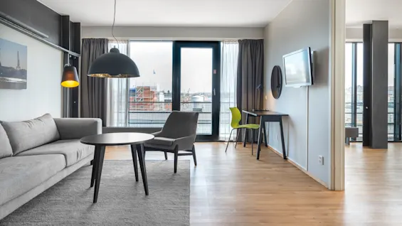 A gray sofa and round table sit on a rug beneath pendant lights in front of large windows with a cityscape view, alongside a desk and chair.  