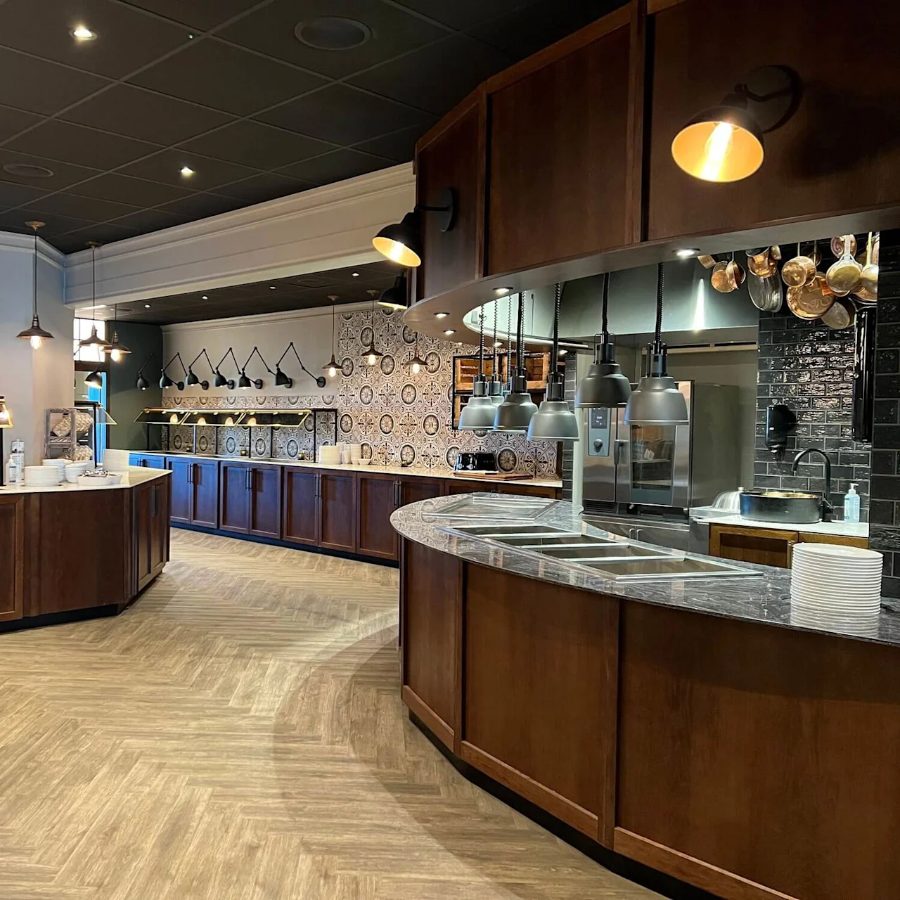 Buffetborde, oplyst af hængelamper, venter på servering i et moderne, varmt dekoreret restaurationsinteriør. Rustfrit stål og dekorative fliser pryder væggene.