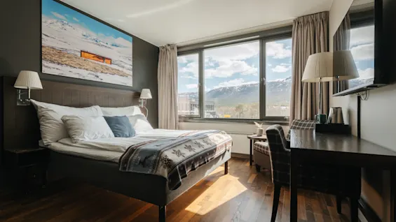 Ljust och modernt hotellrum med dubbelsäng, skrivbord och stora fönster som släpper in naturligt dagsljus.