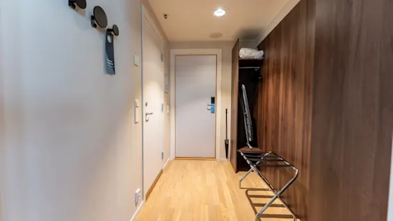 En hotellromsinngang med tregulv, en bagasjehylle og en garderobe. Et "ikke forstyrr"-skilt henger på veggen.