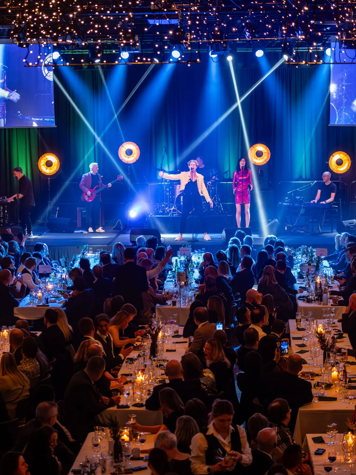 Concert performance, with musicians playing on a stage under bright lights; seated audience fills tables, creating a formal, lively atmosphere. Screens display close-ups of the performers.
