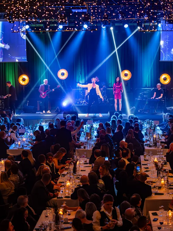Konsertopptreden, med musikere som spiller på en scene under skarpt lys; sittende publikum fyller bordene, og skaper en formell, livlig atmosfære. Skjermer viser nærbilder av artistene.