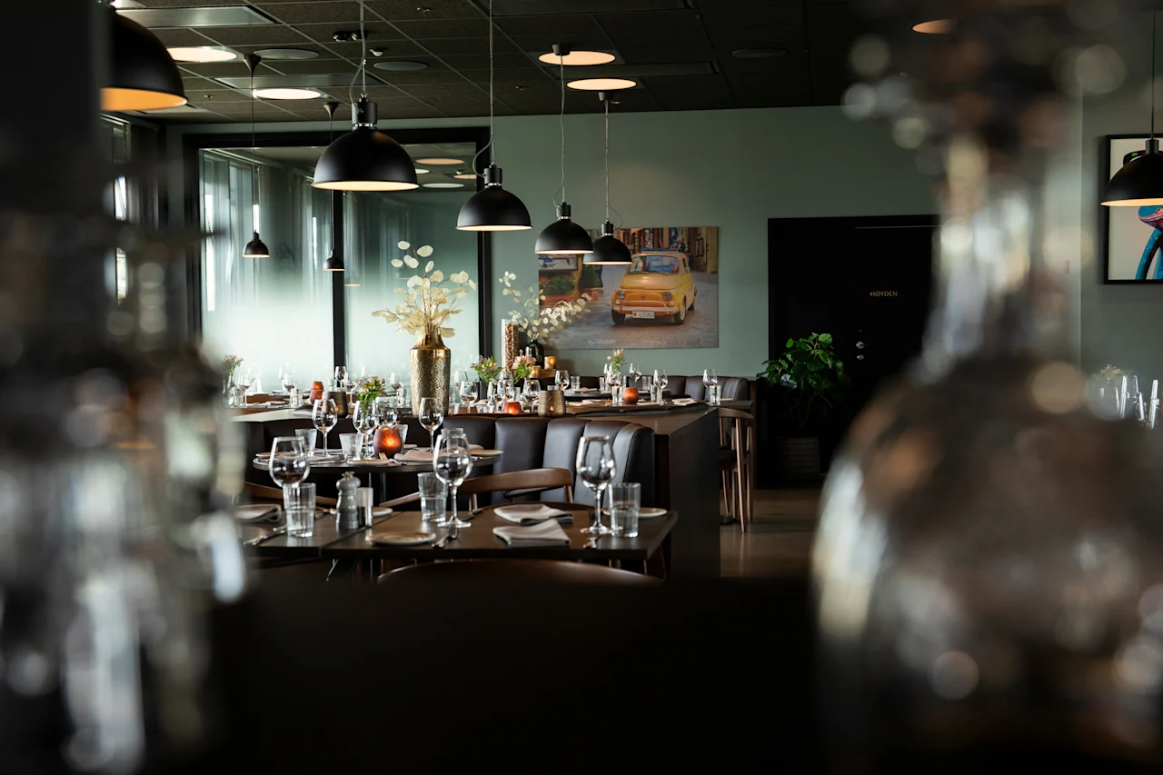 Et moderne restaurantinteriør med dekkede bord, pendellamper og et maleri av en gul bil. En dør med teksten 'HINDEN' er synlig.