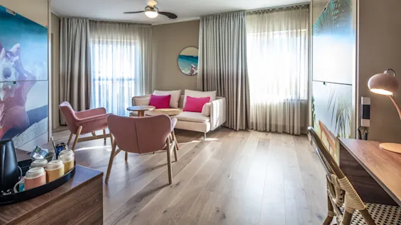 A living room with a white sofa and pink chairs, adorned with pink pillows. Sunlight filters through sheer curtains, highlighting wooden floors. A desk, chair, and wall art complement the cozy space.