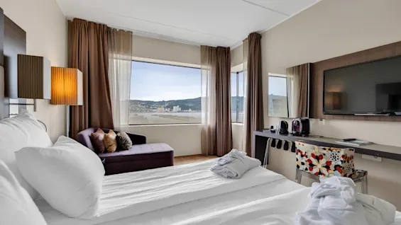A neatly made bed with pillows and robes rests near a purple chaise lounge by large windows, offering a scenic city view. A wall-mounted TV and coffee machine sit on a desk.