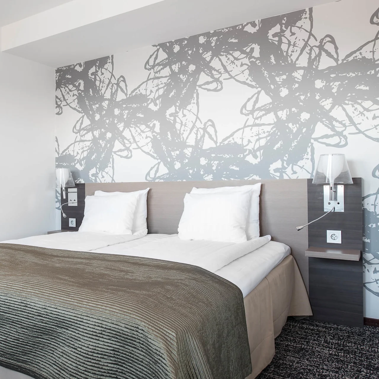 En prydligt bäddad säng med vita kuddar och en brun filt står i ett modernt hotellrum, prytt med abstrakta gråa väggmönster och sänglampor på varje sida.
