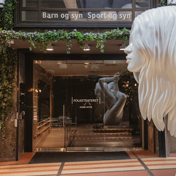 Building entrance with green plants. Inside, a statue in a yoga pose. Text above door: Barn og syn Sport og syn.